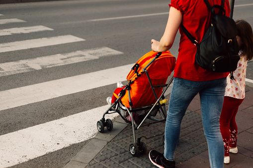 Young mother with her little daughter and baby in stroller waiting on empty crosswalk.