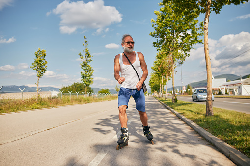 Handsome bearded senior man with sunglasses enjoying a ride on roller skates outdoors in summer on a sunny day, listening to music on headphones.