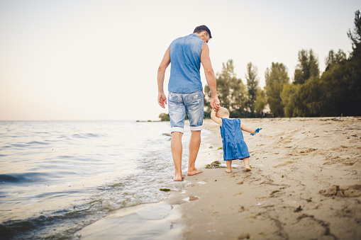 Father and daughter holding hands. female toddler holding fathers hand whilst on a sandy beach by the sea at sunset. Together travel love family Father s day holiday concept. Dad teach child to walk.