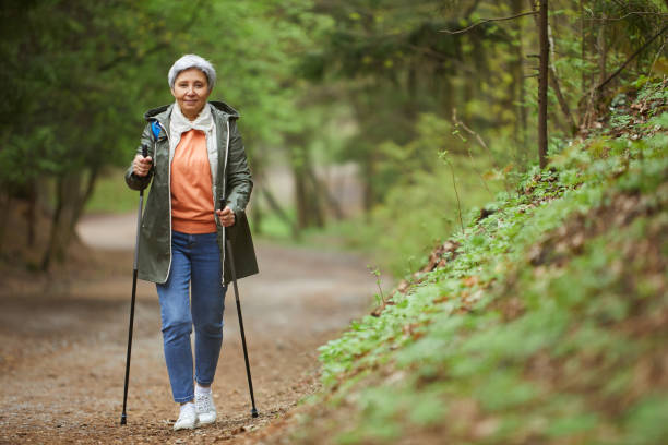 Senior Woman Enjoying Nordic Walk in Forest Full length portrait of active senior woman walking towards camera with Nordic poles while enjoying hike in beautiful autumn forest, copy space nordic walking pole stock pictures, royalty-free photos & images
