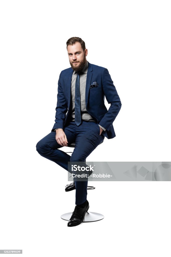 Attractive Caucasian businessman wearing an elegant blue suit and sitting on a stool Confident, attractive and
bearded CEO looking elegant in his custom made blue suit and
tie, with one hand in his pocket and sitting on a stool, isolated on
white background. Sitting Stock Photo