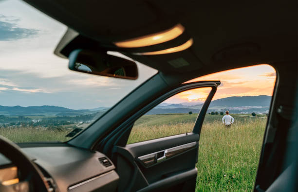 jeune homme restant dans l’herbe élevée et appréciant les couleurs de ciel de coucher du soleil à travers la vue ouverte de dor de voiture. voyager en voiture concept image. - car road highway open photos et images de collection