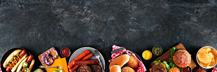 Summer BBQ food table scene with hot dog and hamburger buffet. Overhead view bottom border over a dark slate background. Copy space.