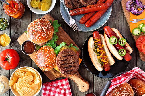 Summer BBQ food table scene with hot dog and hamburger buffet. Top view over a dark wood background.