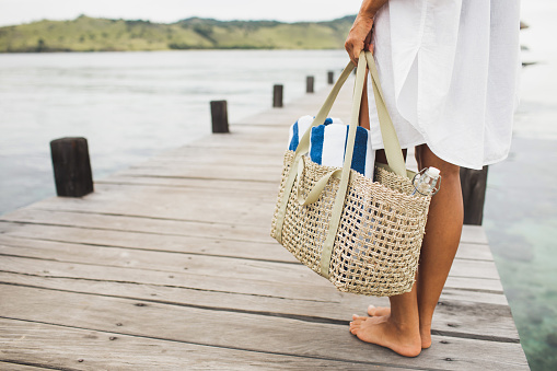 Woman holding straw wicker bag with two beach towels and glass bottle for water and going to the beach. Eco friendly and zero waste concept.