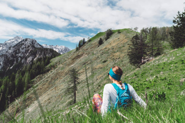 Young woman with sunglasses, wearing  a blue backpack, hiking in the mountains on a beautiful day, taking a stop to enjoy the views, sitting on the grass.. Adventurous woman hiking in the Alps alone. Scenic views on the mountains.