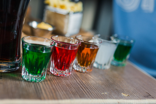 Multi-color cool alcohol party drinks with on wooden table in bar. Bright colorful glasses with alcoholic shots. Mixed liquor, vodka and juice in shot glasses. Alcoholic cocktail row on bar table