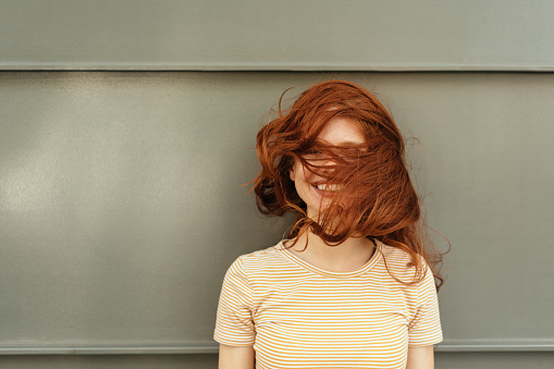 Happy smiling woman with her red hair covering her face standing in front of a grey wall in a head and shoulders portrait