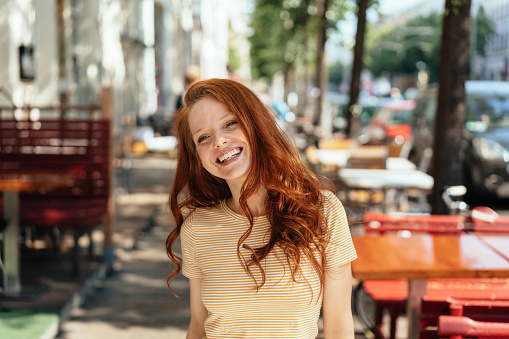 Attractive young redhead woman with a lovely warm friendly smile standing at an outdoor restaurant in town grinning at the camera