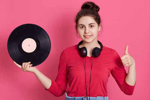 Young adorable girl showing thumb up and holding vinyl music plate in hand, female with knot and head phones around neck looking at camera, listening retro music via gramophone.