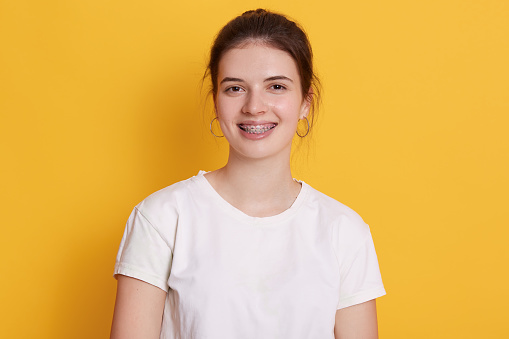 Mujer sonriente con soportes y pendientes redondeados posando contra la pared amarilla, mujer con pelo oscuro mirando a la cámara con expresión facial feliz, usando camiseta blanca. photo