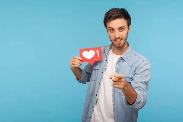 Photo of Hey you, click Like! Portrait of handsome blogger man in denim shirt holding social media heart Like button