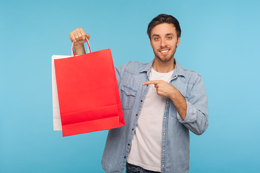 Portrait of stylish handsome brunette man in denim shirt pointing red shopping bags and smiling, showing packages with purchase from fashion store. indoor studio shot isolated on blue background