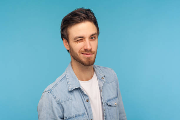 Portrait of cheerful handsome man in worker denim shirt winking to camera, blinking eye joyfully Portrait of cheerful handsome man in worker denim shirt winking to camera, blinking eye joyfully as if having interesting idea, looking playfully flirting. studio shot isolated on blue background young man wink stock pictures, royalty-free photos & images