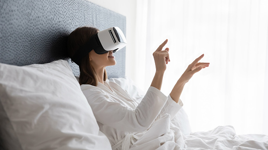 Amazed young woman sit wake up in white bed wearing virtual reality glasses, stunned millennial girl awaken rest in hotel or home bedroom try experience VR googles, modern technology concept