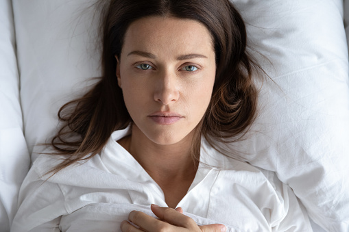 Top view of depressed upset young woman lie relax on white pillow linen sheets feel distressed lonely, sad millennial female rest in home bedroom suffer from depression or insomnia, having problems