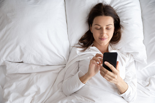 Top view of smiling young woman awaken in white cozy bed using modern smartphone gadget, happy millennial female wake up lie in comfortable bedroom browsing Internet on cellphone in the morning