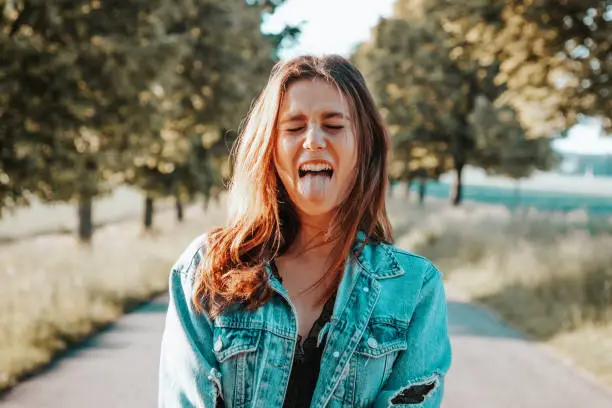 Young woman standing in the middle of country road making fun, grimacing, sticking out tongue, playing crazy. Millennial Generation Youth Summer Lifestyle Real People.