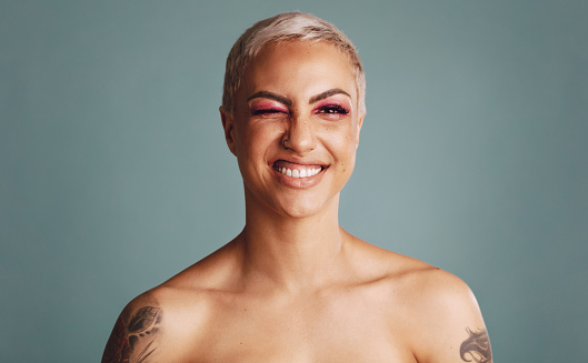 Smiling woman looking at camera and winking an eye. Beautiful female model with blonde pixie hairstyle having tattoo on her hands standing on grey background.