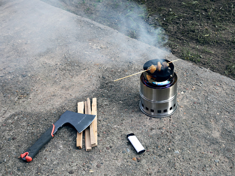 Portable wood burning stove smokes meat on a wooden stick. Nearby lies an ax for splitting firewood slivers