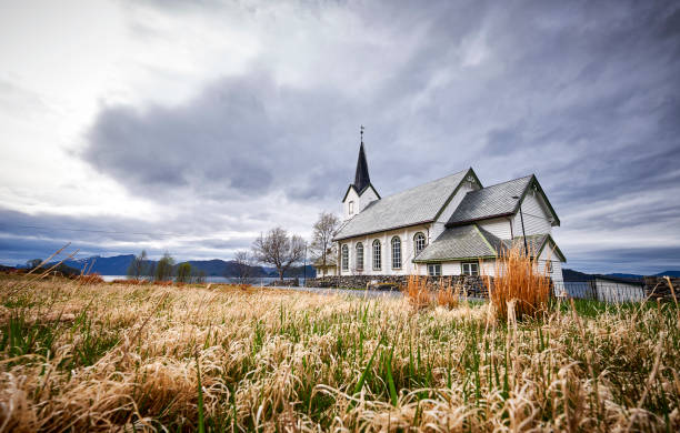A white church in Norway, it’s located near a lake and surrounded by different types of grass and some trees.