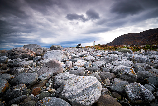 In the foreground you see many big boulders, in the background there’s a car and the lighthouse of Alnes in Norway.