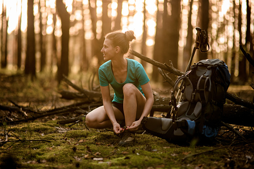 young woman in forest ties shoelaces on shoes and looks away. Next to her lies sports backpack and sticks