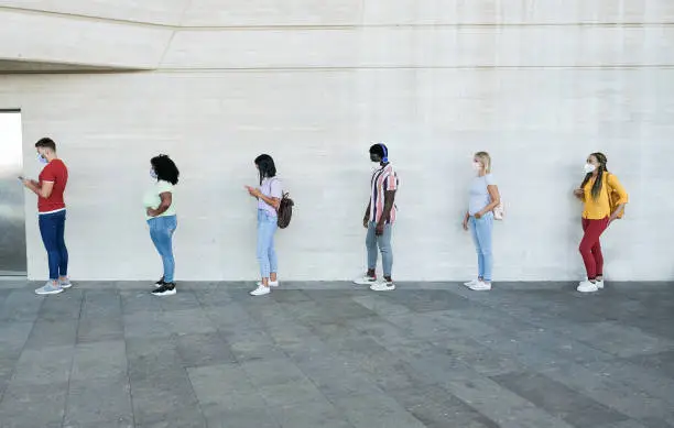 Photo of Multiracial people standing in a queue and waiting - Young people with social distancing and wearing protective face masks - Concept of the new normality and social distancing