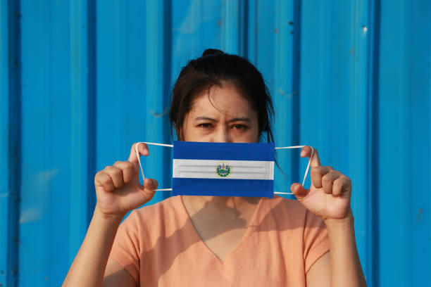 una mujer con bandera de el salvador en máscara higiénica en la mano y levantó la cara frontal sobre fondo azul. - arms lifted fotografías e imágenes de stock