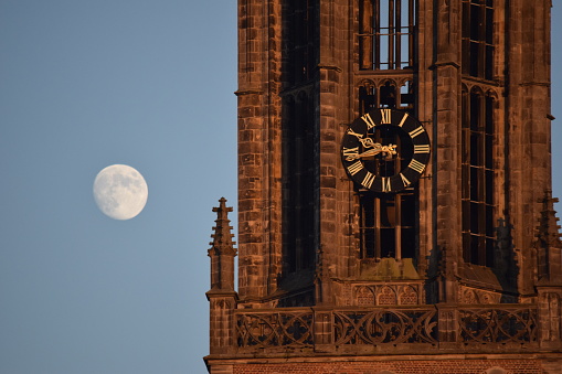 It is almost ten o'clock in the evening when the sunset shines on the clock tower of Amersfoort with the moon on the background, during the Dutch summertime period.