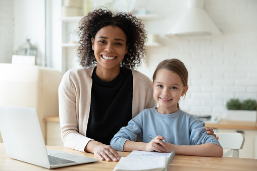 Happy African female tutor and little learner schoolgirl portrait. Multi-racial family sit in kitchen laptop and workbooks lie on table. Successful homeschooling, clever kid girl study at home concept