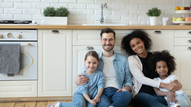 Multiracial couple with adorable daughters sit on floor in kitchen Horizontal photo multiracial excited couple with adorable daughters sit in modern renovated kitchen on warm floor smile look at camera. New property owners, happy multicultural family portrait concept adoption photos stock pictures, royalty-free photos & images