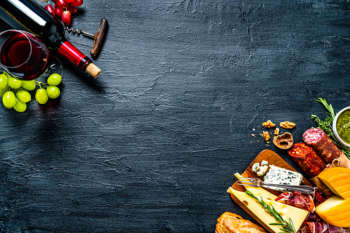 Appetizer backgrounds: delicious traditional Spanish tapas shot from above on dark background. The arrangement includes red wine, Spanish chorizo, various cheeses, Serrano ham and bread. The composition is at the top left and bottom right of an horizontal frame leaving useful copy space for text and/or logo at the center. High resolution 42Mp studio digital capture taken with SONY A7rII and Zeiss Batis 40mm F2.0 CF lens