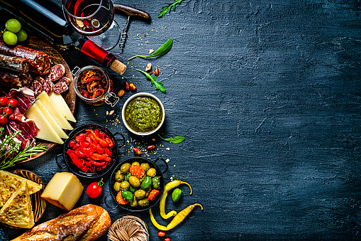 Appetizer backgrounds: delicious traditional Spanish tapas shot from above on dark background. The arrangement includes red wine, Spanish chorizo, various cheeses, Serrano ham, Spanish omelet, olives and pickles, sun dried tomatoes, jalapeño pepper and bread. The composition is at the left of an horizontal frame leaving useful copy space for text and/or logo at the right. High resolution 42Mp studio digital capture taken with SONY A7rII and Zeiss Batis 40mm F2.0 CF lens