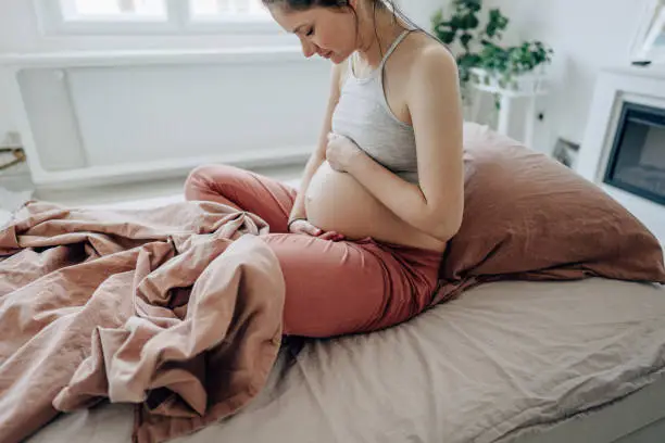 Photo of a young expectant mother stroking her baby bump and enjoying her morning in the bedroom of her apartment; the daily routine of a pregnant woman.