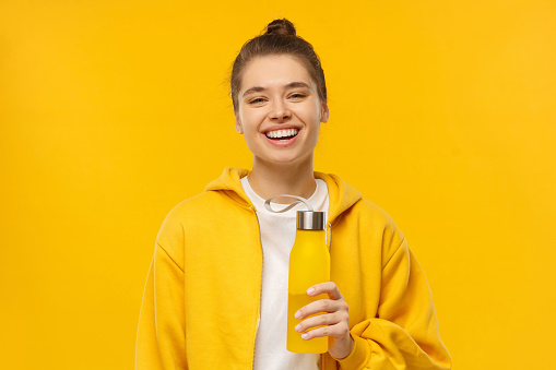 Young sports woman in hoodie and white t-shirt, holding reusable eco water bottle, isolated on yellow background