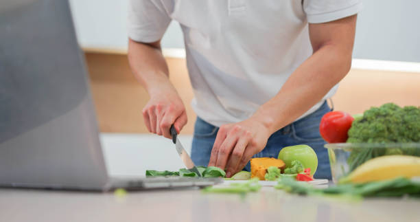 close up man hand using knife to cut vegetables on board and preparing food while open laptop to practice from streaming website and cooking online course for healthy living concept