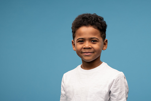 Happy cute little boy of African ethnicity in white t-shirt standing in front of camera against blue background in isolation