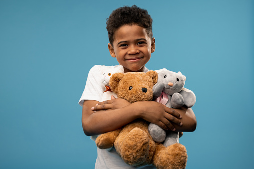 Happy cute little boy with two soft toys and teddybear smiling at you while playing in studio in front of camera on blue background