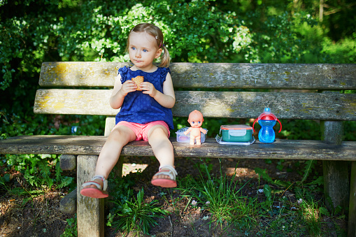 Adorable toddler girl sitting on the bench and having picnic on a summer day. Child enjoying healthy snack in park. Outdoor summer activity for small kids
