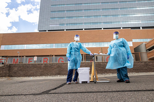 Two Female Nurses Wearing Protective Gowns, Surgical Face Masks, Gloves, and Face Shields Look at the Camera while Waiting for Patients in a Drive-Up (Drive-Through) COVID-19 (Coronavirus) Testing Line Outside a Medical Clinic/Hospital Outdoors