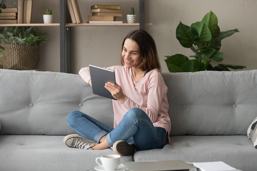Young smiling woman sitting on couch, using tablet, watching funny video or moving, chatting with friends, reading email with good news, web surfing, holding video call or posting in social networks.