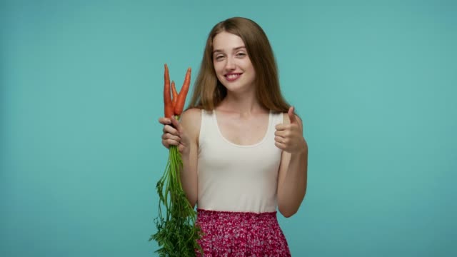 Positive cute girl in summer dress smelling raw carrot and showing thumbs up, like gesture, enjoying fresh vegetables