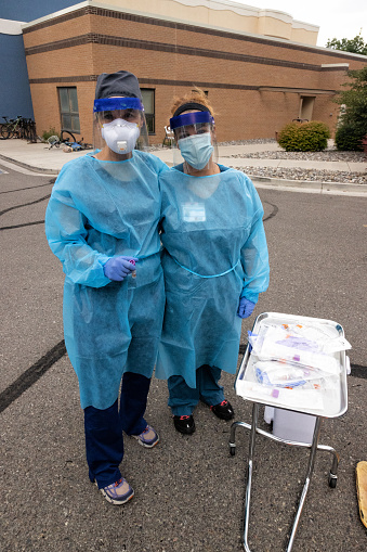 Two Female Nurses Smile While Wearing Gowns, Surgical Face Masks, Gloves, and Face Shields Look at the Camera while Waiting for Patients in a Drive-Up (Drive-Through) COVID-19 (Coronavirus) Testing Line Outside a Medical Clinic/Hospital Outdoors