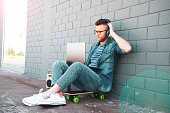 Young man with headphones using laptop in the street - Trendy cool guy having fun surfing online and listening music - Influencer or blogger working outdoors - Technology and fashion lifestyle concept