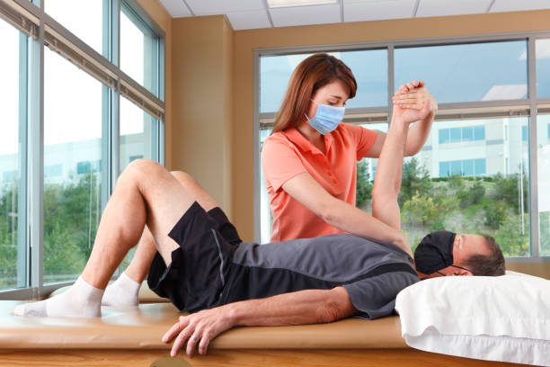 Physical Therapist Performing Shoulder PNF Pattern On Male Patient While Both Wear Protective Masks A female physical therapist performs a shoulder PNF pattern on a male patient as he lies on a treatment table in a clinical setting.  Both the therapist and the patient wear protective masks to prevent the spread of COVID-19 or other infectious diseases. The therapist is in her early thirties and the patient is in his mid 50's. physical therapist photos stock pictures, royalty-free photos & images