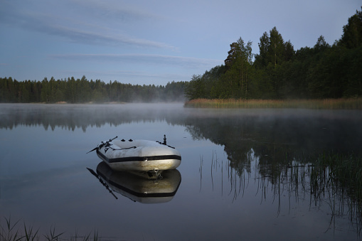 Fog is curling over the grassed lake banks at dawn, an inflatable boat is in the foreground, Tver region, Russia