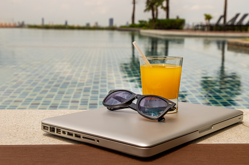 Relaxing moment by the pool. Take a break from working on laptop and enjoy the fresh orange juice for a refreshment.  Outdoor swimming pool background. leisure activity.