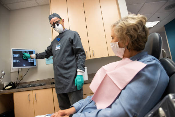 un dentista maschio che indossa una maschera n95, guanti chirurgici e loupes discute i raggi x dei denti su uno schermo del computer con una paziente mascherata sulla sess anni sessanta in una sala d'esame in una clinica dentale - dentist dentist office patient discussion foto e immagini stock