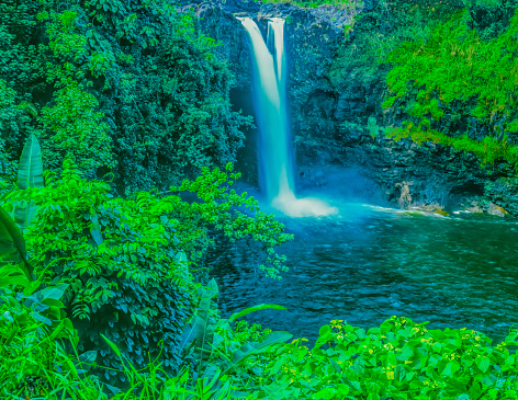 Lush and fresh vegetation surrounds Rainbow Falls near Hilo, Hawaii. The waterfalls has twin falls at the top and they both pour into a pond below.